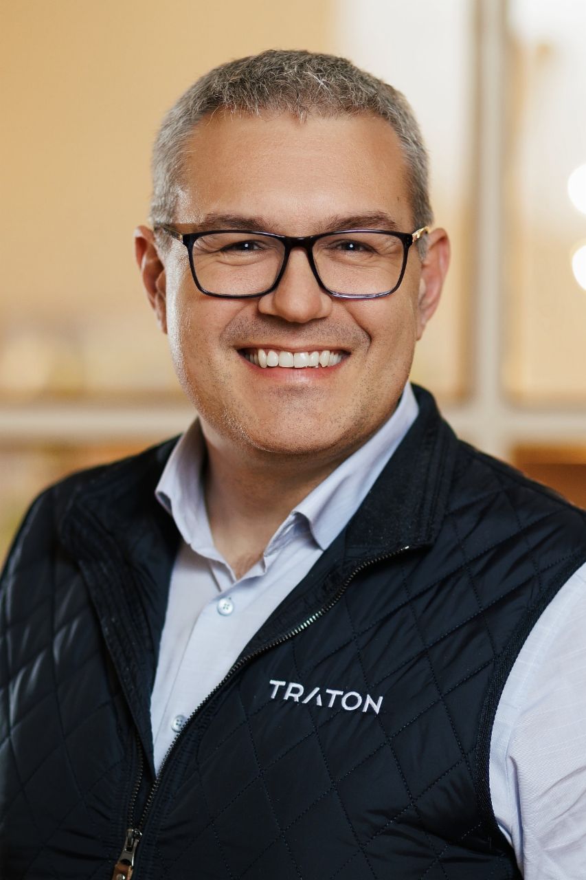 Stefan Riegl, Bereichsleiter Systems Engineering bei TRATON GROUP R&D, trägt eine schwarze Brille, ein blaues Hemd und eine schwarze Weste mit dem TRATON-Logo auf der rechten Seite. 