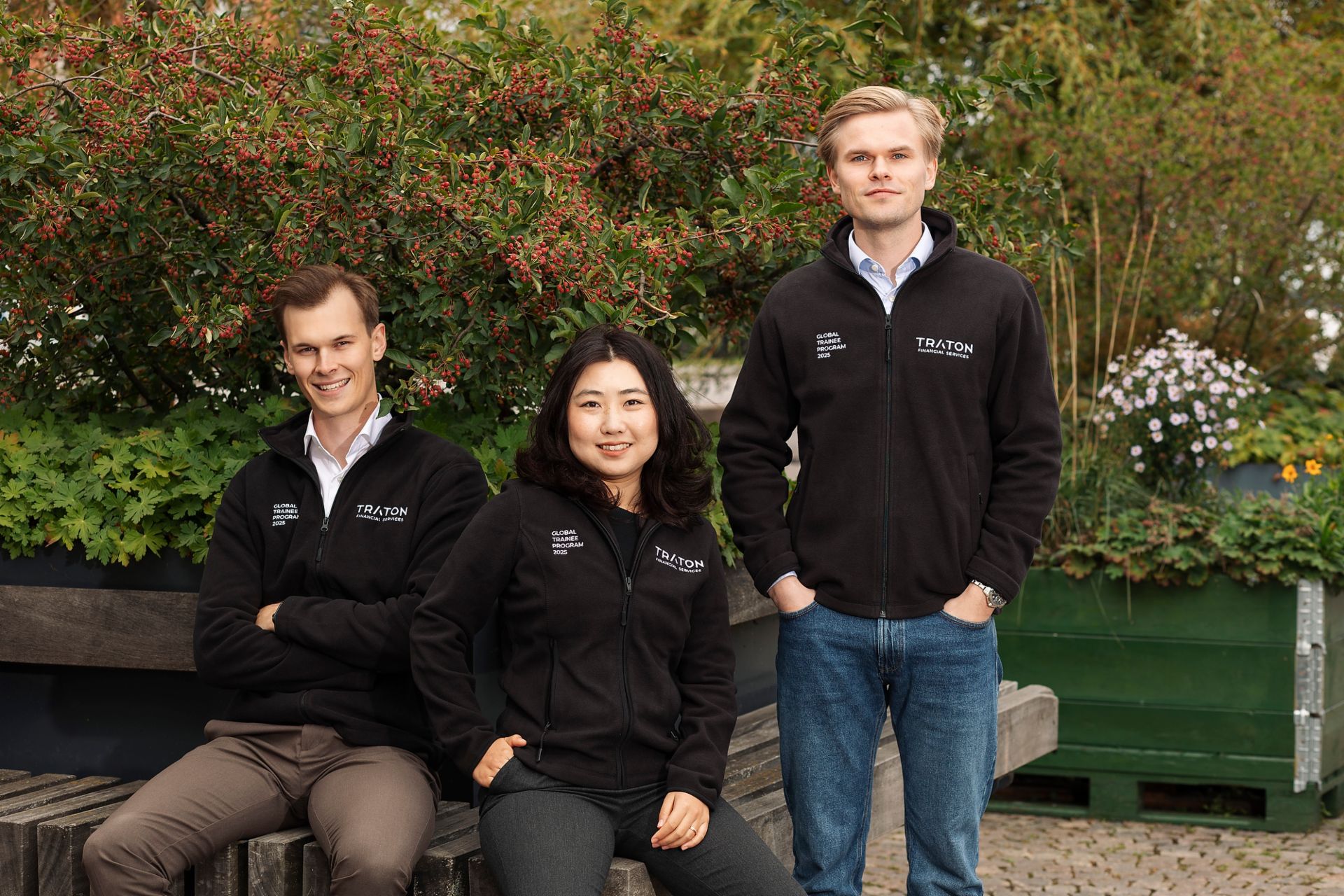 Drei junge Leute, die einen Pullover mit der Aufschrift „Global Trainee Program“ von TRATON Financial Services tragen. 
