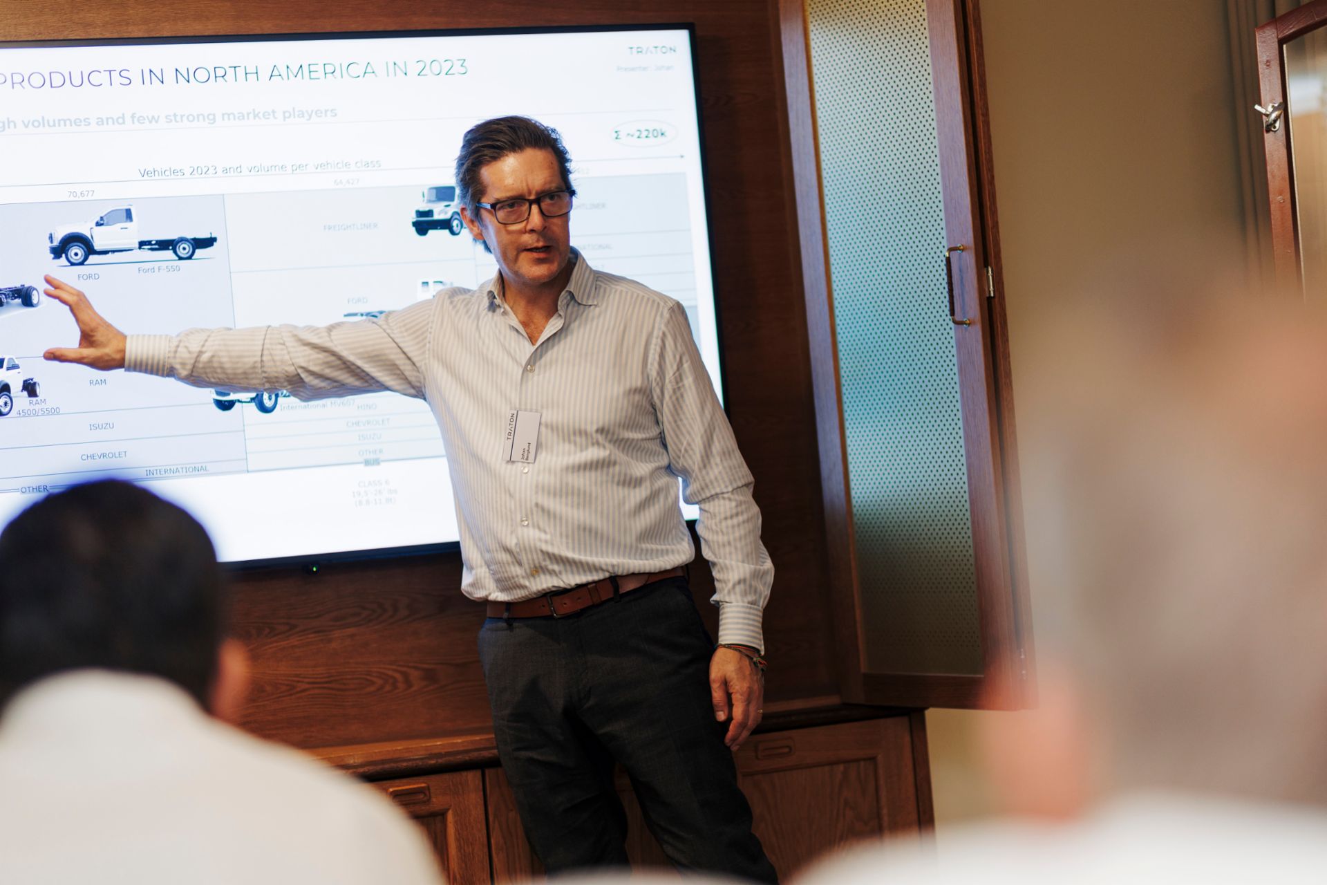 Johan Berglund, Strategic Product Planner at TRATON GROUP, wearing a white shirt with blue stripes. He is pointing at a screen in which he is presenting 2023 vehicles and volume per vehicle class. 