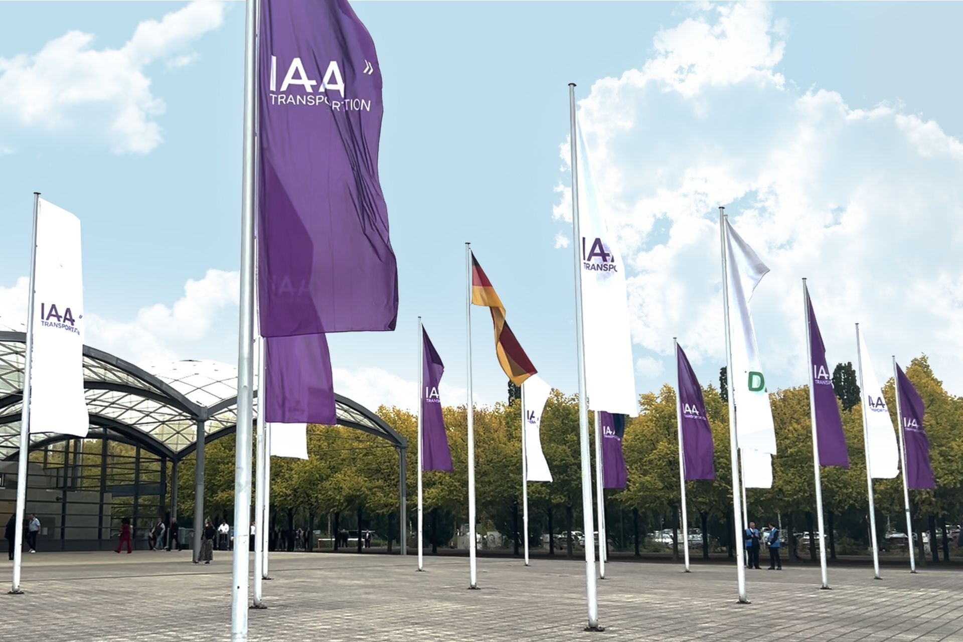 Ein Foto, das mehrere Fahnen mit dem IAA Transportation-Logo zeigt, die vor einer modernen Ausstellungshalle wehen. Die Fahnen sind lila und weiß, eine deutsche Fahne ist ebenfalls sichtbar. Im Hintergrund sind ein teilweise bewölkter Himmel und Bäume mit grünem Laub zu sehen. Menschen gehen und versammeln sich vor dem Eingang der Halle.